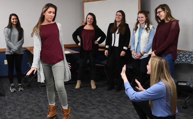 During an improv workshop, one young woman plays a photographer and another plays a model, while several other students look on. 