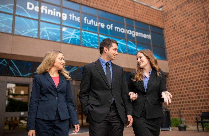 Three MBA students walk away from Jacobs Management Center, with the words "Defining the future of management" visible on the building behind them. 