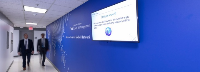 Two students walk by the Office of Alumni Engagement and External Relations; the wall beside them features a world map and digital screen. 