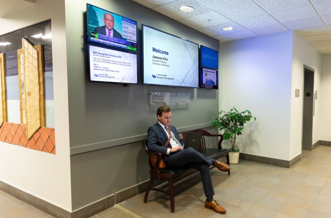 A student works on his laptop; on the wall behind him is a large flatscreen with a welcome message and four smaller television screens. 