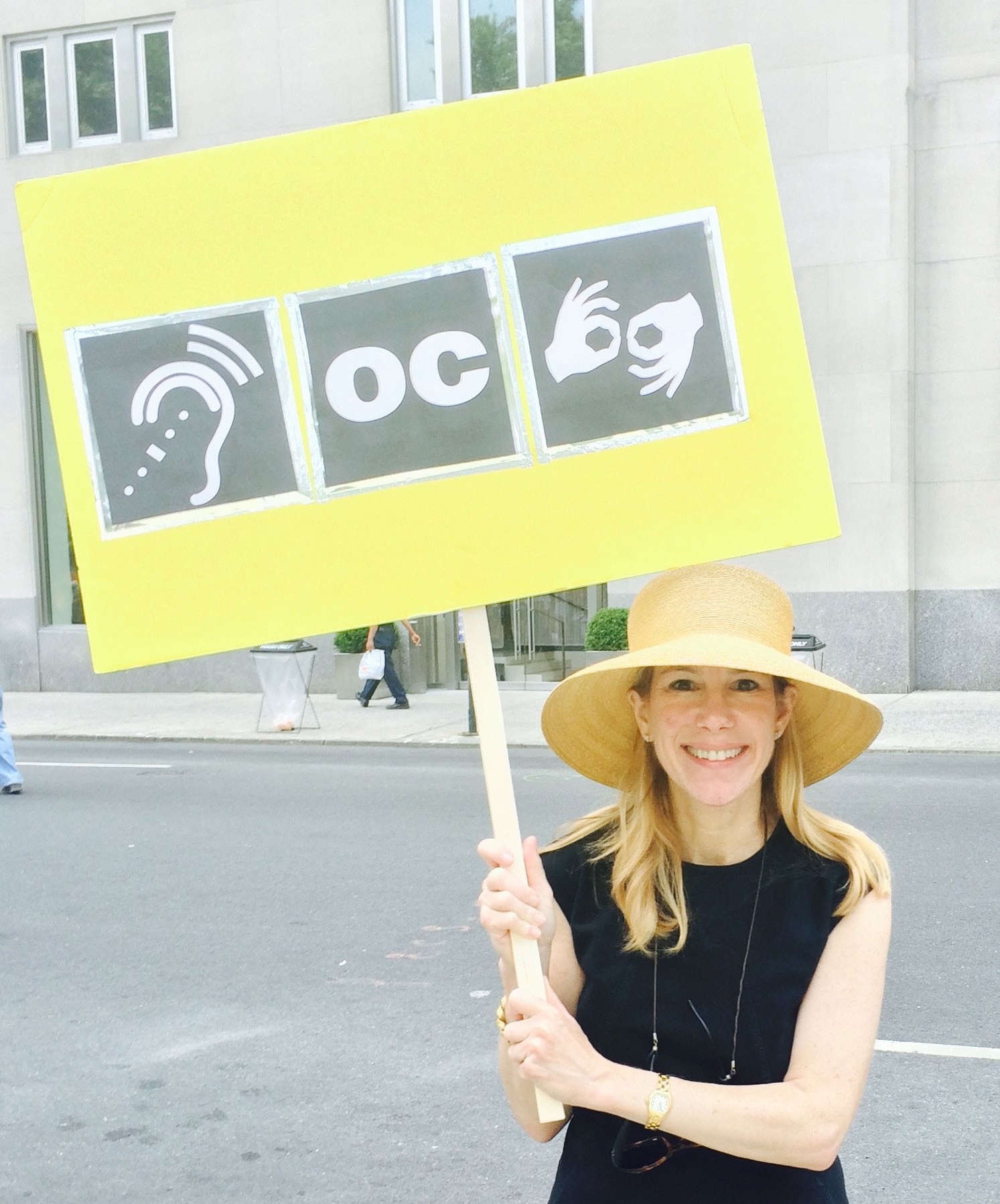 Zoom image: Lintz holds a sign with symbols that indicate the presence of critical accessibility services for people with hearing loss: assisted listening devices, open captioning and sign language interpretation. 