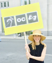 Janice Lintz holds a sign with three symbols for assisted listening devices, open captioning and sign language interpretation. 