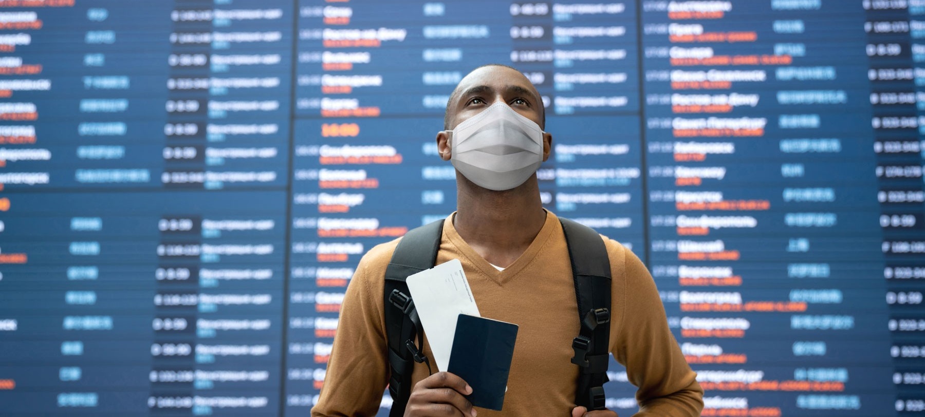 Man wearing a mask stands in front of the departures board. 