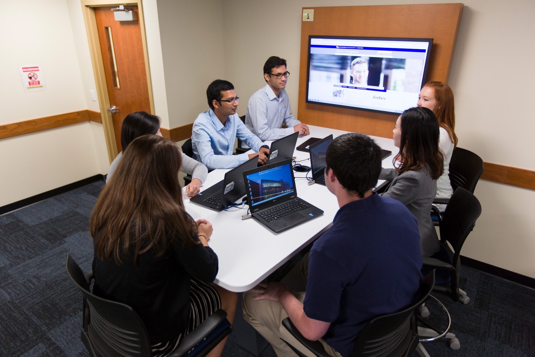 Students sitting discussing in the new undergraduate center on laptops and an LED display screen. 