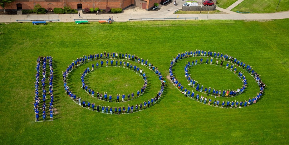 People standing on a grassy field in the shape of the number 100. 