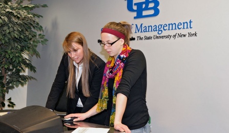 Two students at a podium looking at the computer in the podium. 