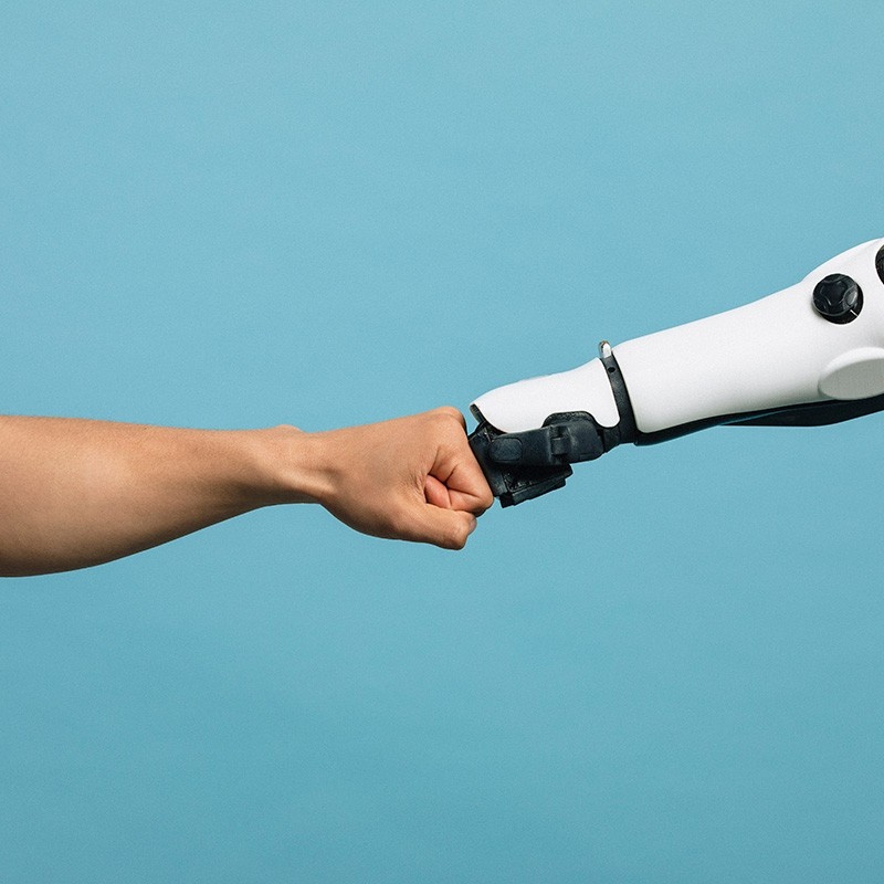 Photo of a human arm fist bumping a robotic arm. 