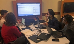Students with laptops gathered around a larger screen, collaborating on a database project. 