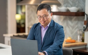 School of Management graduate student working on a laptop at home in a kitchen. Link goes to MS Online Business Analytics homepage. 