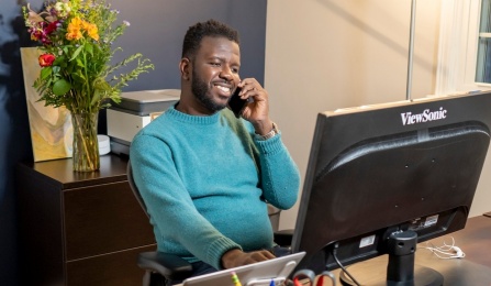 School of Management graduate student talking on a call phone sitting in a home office. Link goes to Online MBA homepage. 