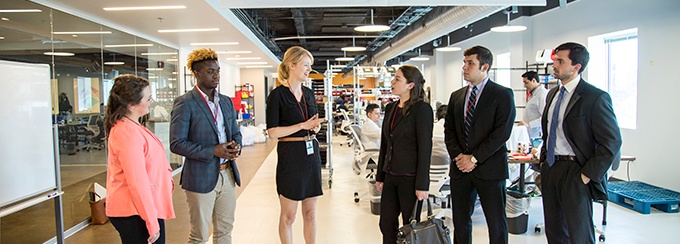 Five UB MBA students standing in a work environment listening to woman in between them talking.