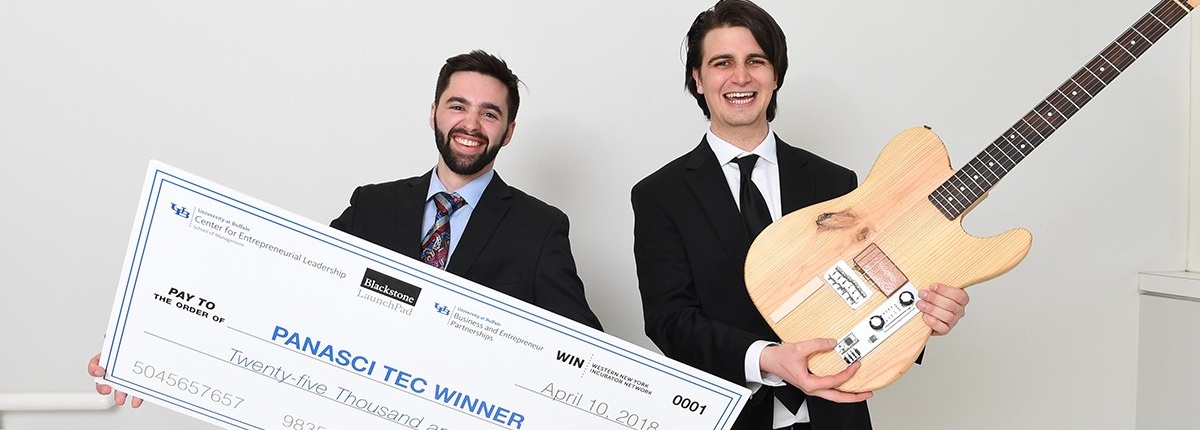Two male students in professional business attire. Student on the left of photo is holding a large $25,000 check. The student on the right is holding a guitar. 