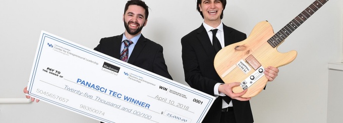 Two male students in professional business attire. Student on the left of photo is holding a large $25,000 check. The student on the right is holding a guitar. 