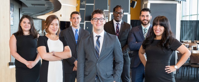 Seven MBA students dressed in professional business attire standing and looking at the camera. 