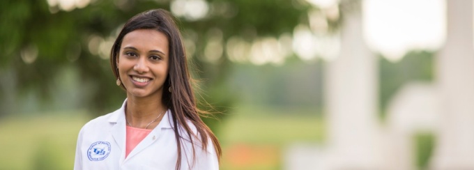 Female medical student in lab coat standing in left side of photo with trees in background. 