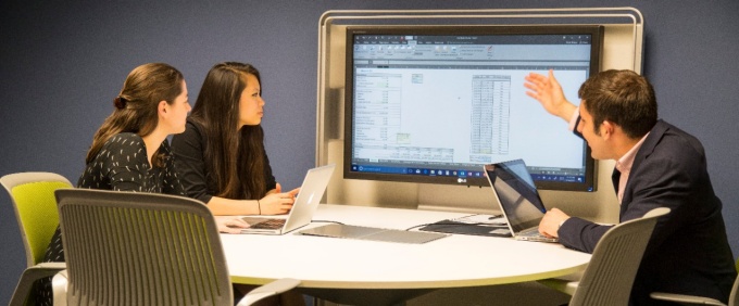 Three MBA students sitting at a table with a large monitor at the end of the table. Two female students are on the left of the photo and one male student on the right is pointing at the large monitor. 