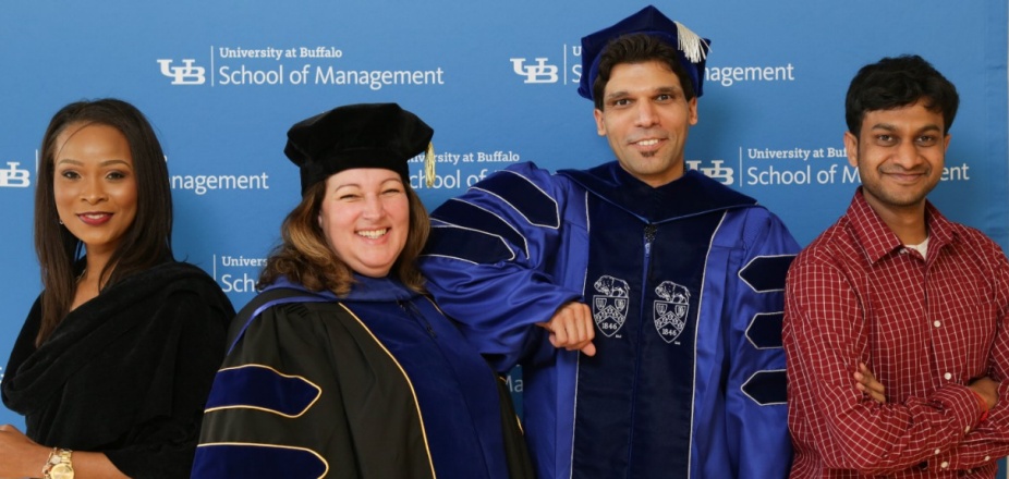 A group of four PhD in Management graduates, two of them in caps and gowns. 